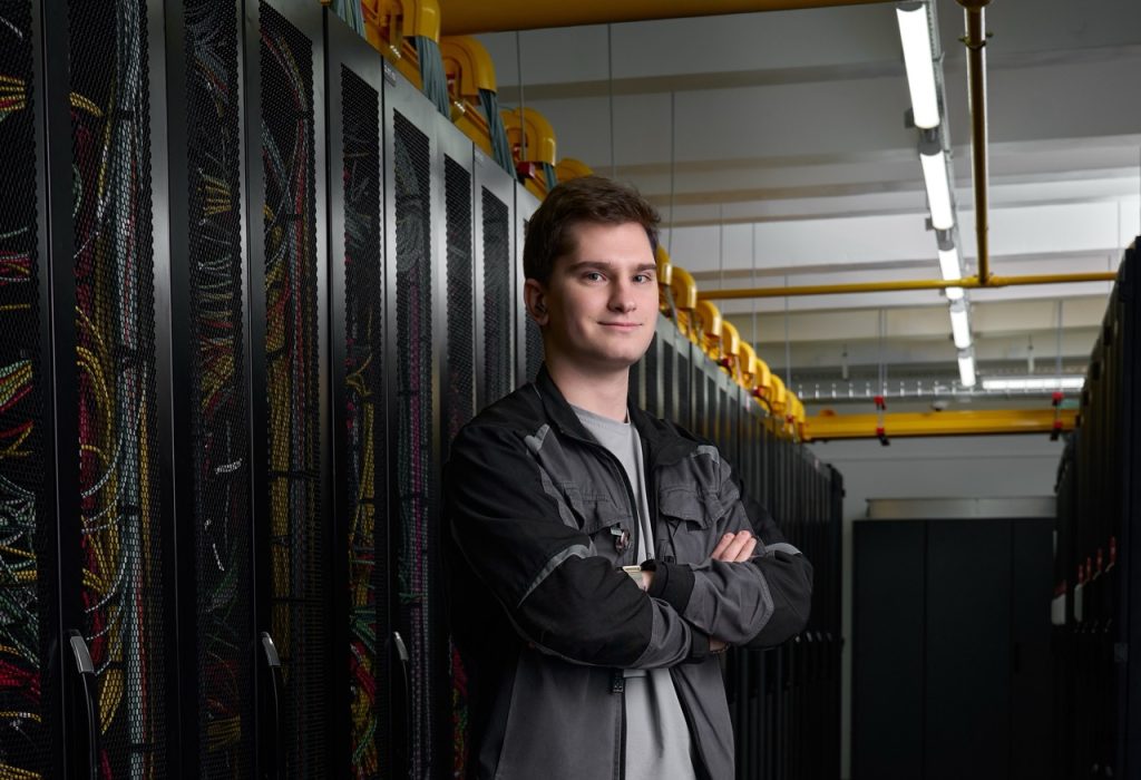 Young technician standing with arms crossed in a server room beside row of rack cabinets and tangled cables.