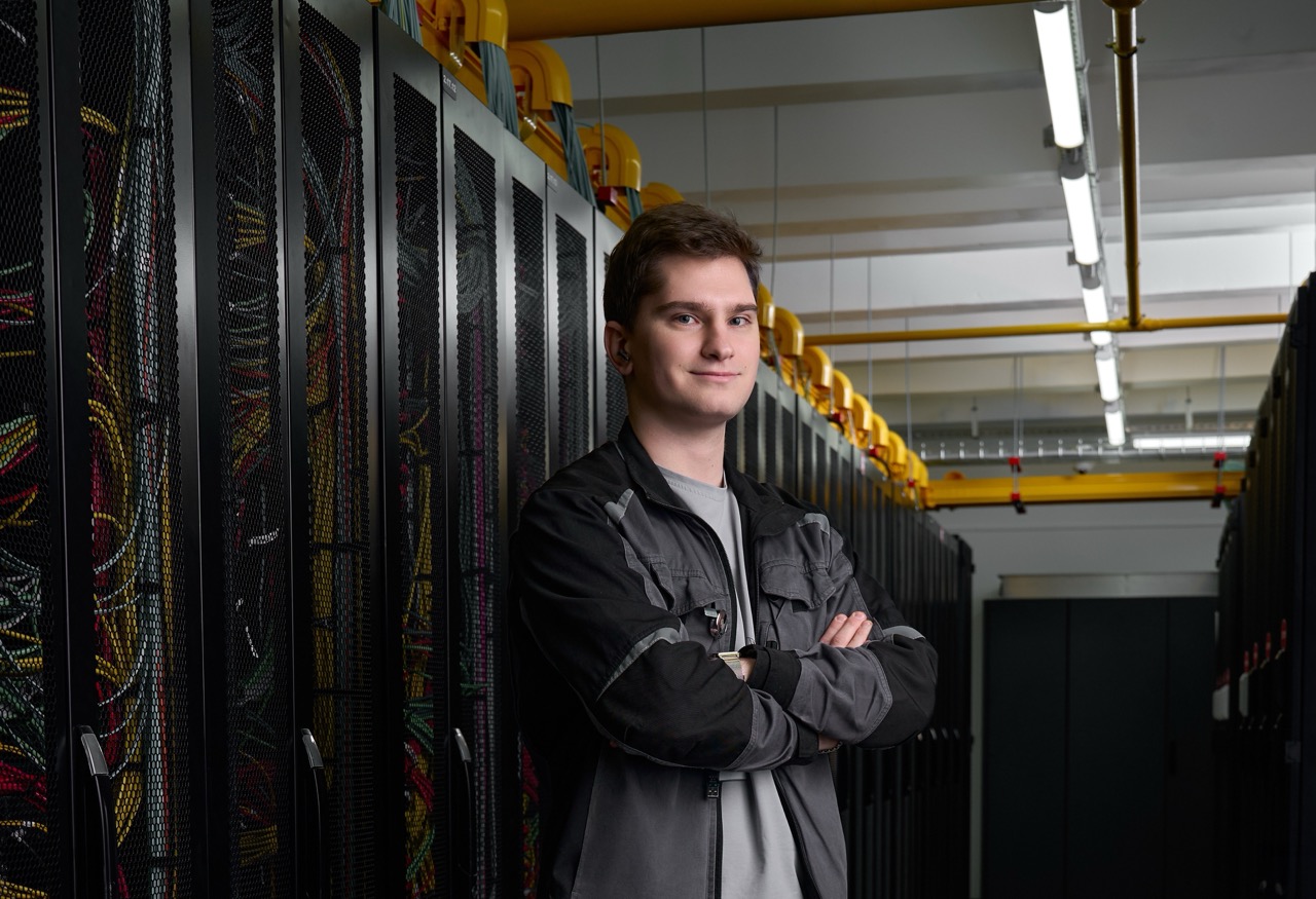 Young technician standing with arms crossed in a server room beside row of rack cabinets and tangled cables.
