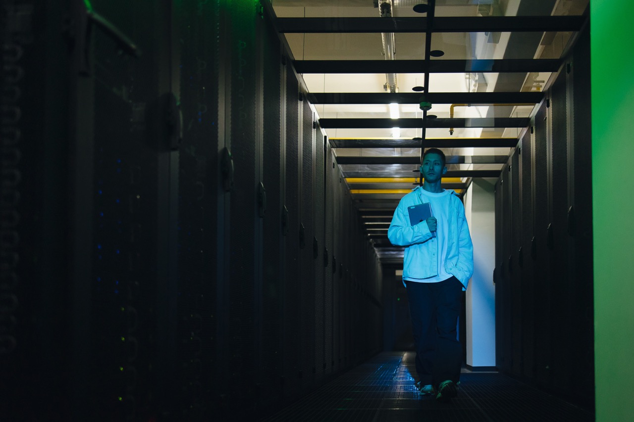 Person in a light jacket carrying a notebook walks down a dim data center corridor lined with server racks under blue-green lighting.