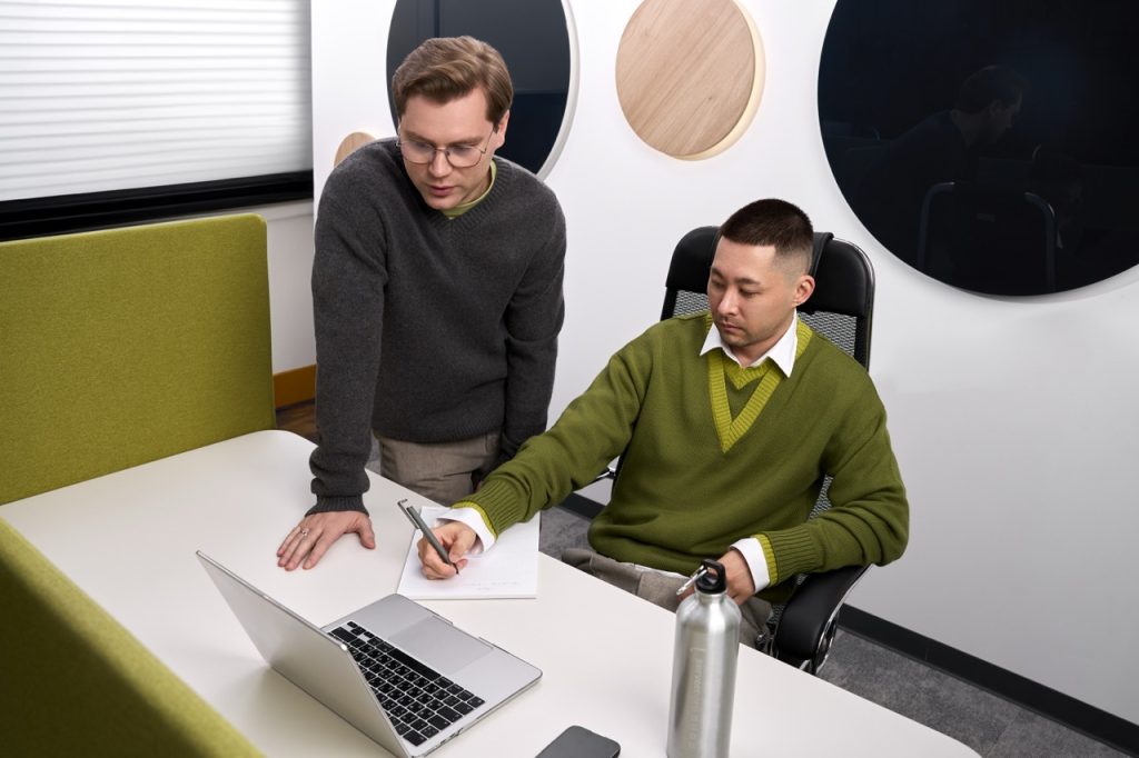 Two coworkers collaborate at a white table: one seated and writing in a notebook while the other leans over to review papers near a laptop and water bottle in a modern office setting.
