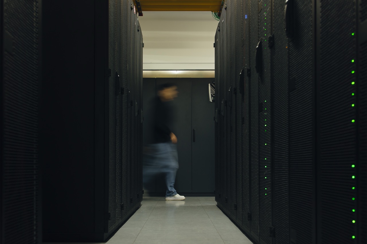 Data center aisle with tall server racks on both sides; a blurred person walks toward a dark door in the distance, green indicator lights visible on the racks.”] } ? Wait format: It should be array of strings. Provide multiple items. I'll craft properly.