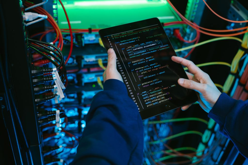 Person holding a tablet displaying Python code in a server rack filled with colorful cables and lights, a tech scene.