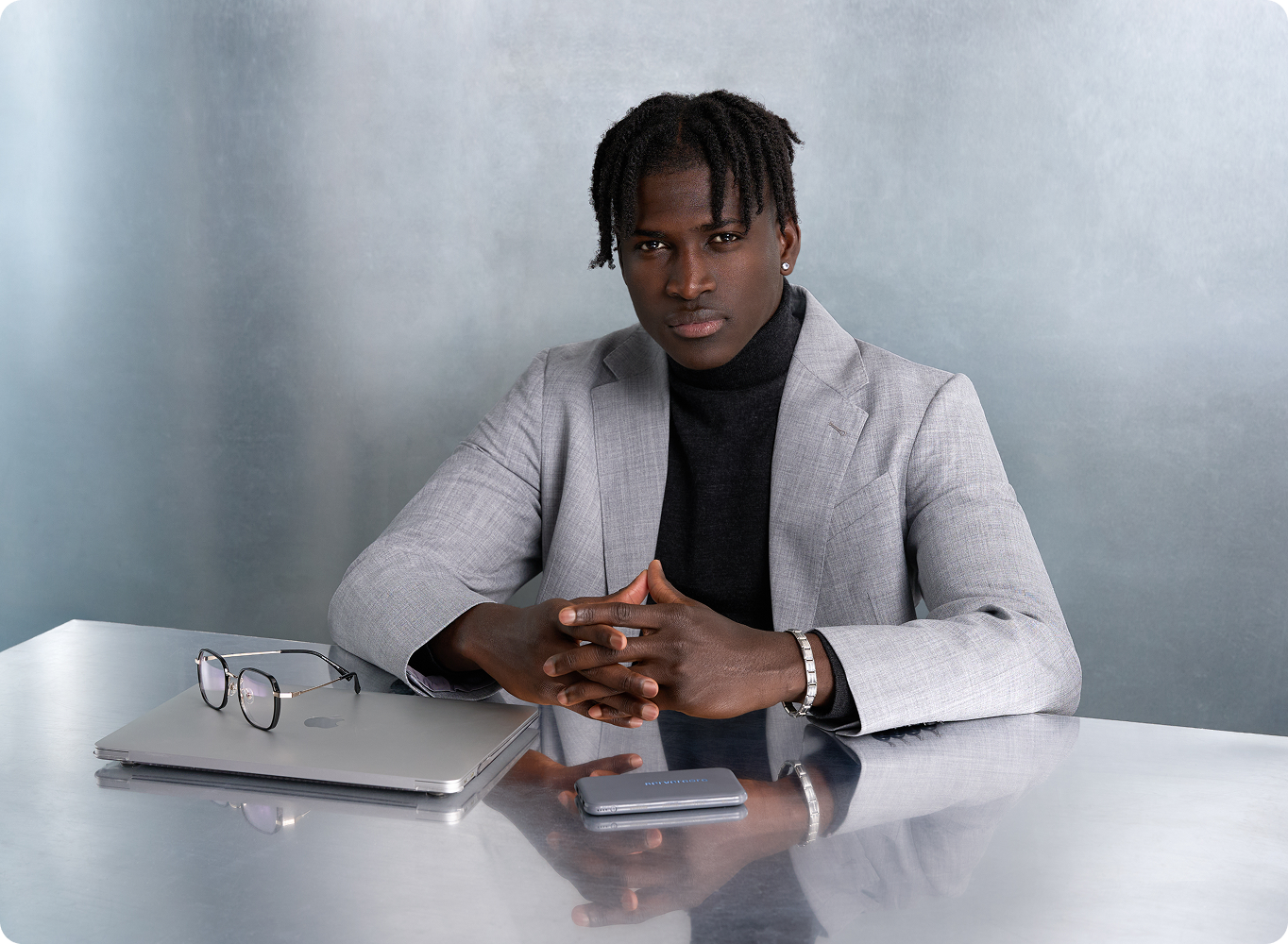 Professional portrait: a man in a gray blazer with a black turtleneck sits at a metallic desk, hands clasped.