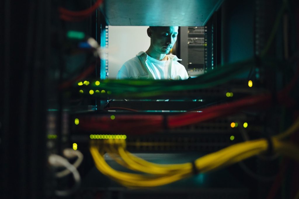 A technician in a white shirt studies a server rack in a dim data center, with colorful cables in the foreground and glowing LEDs nearby.
