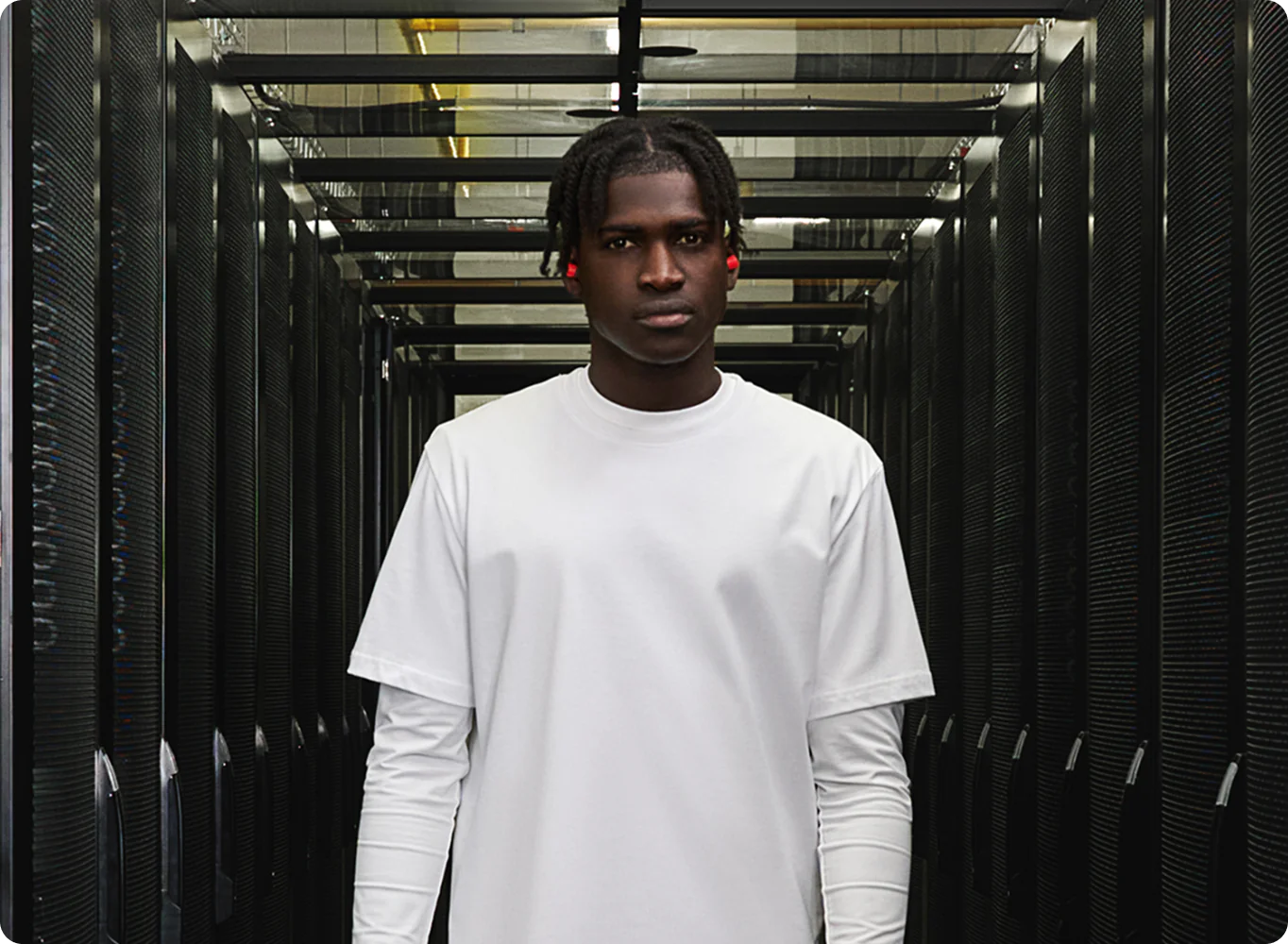 Young man in a white T-shirt standing in a data center hallway between rows of black server racks, looking at the camera.