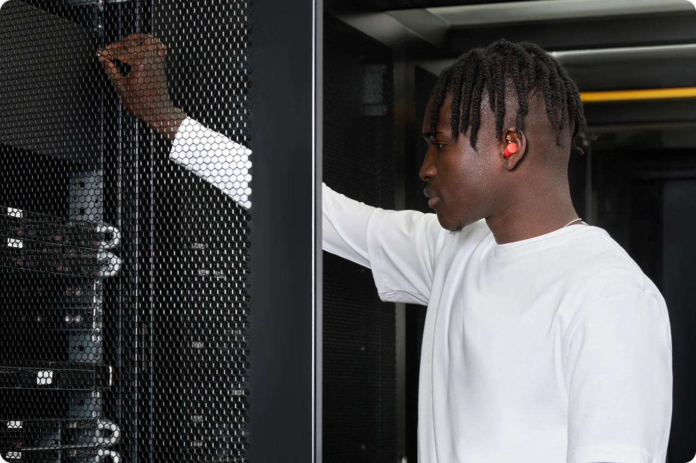 A man in a white T‑shirt works at a server rack, gripping the mesh door while examining networking equipment, wearing red earplugs on his ear.